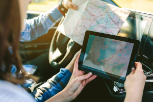 Paar sitzt im Auto und schaut sowohl auf einem Tablet als auch auf einer Karte nach dem Weg.
